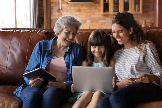 Happy Three Generations Of Latino Women Sit Relax On Couch At Home Using Modern Electronic Gadgets Together. Smiling Little Hispanic Girl With Mom And Grandmother Rest Indoors Have Fun With Devices.