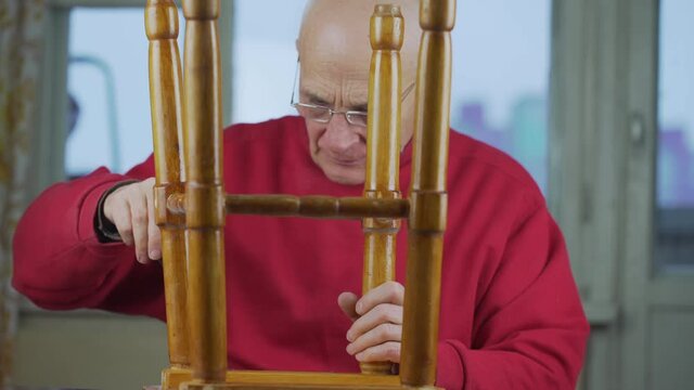 Old man wearing glasses and red sweater repairing wooden stool. Concept household and maintenance.