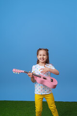 cheerful little Caucasian girl of 4 years old in fashionable bright clothes plays a pink ukulele guitar. The child plays a musical instrument and sings on a lilac background and green grass.