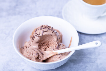 Chocolate ice cream with chocolate sprinkle in a white bowl on a light background. Three balls of homemade ice cream and a cup of coffee. Close-up.