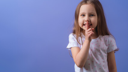 Fototapeta premium portrait of a cute little girl, holding her index finger to her mouth, stands on a purple background. The child doesn't want to talk. Keeps a secret, learned a secret