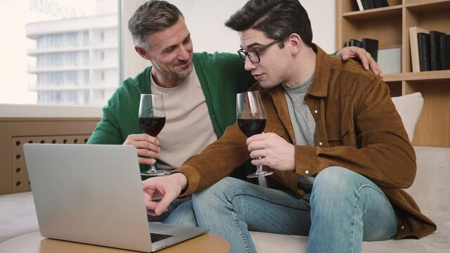 Pleased gay couple are watching laptop while drinking wine and talking sitting in the couch at home