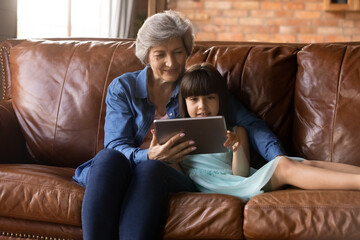 Loving mature Hispanic grandmother relax on sofa in living room with small granddaughter using tablet together. Senior Latino grandparent rest on couch at home with small girl child with pad device.