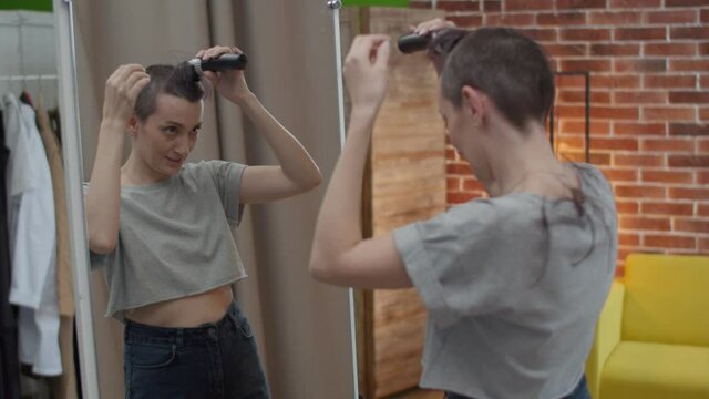 Woman shaves her head bald with a hair machine. The woman is happy to change appearance with a new haircut at the mirror.