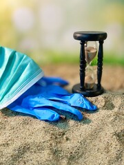 A mask and protective gloves are lying on the sand, the hourglass is counting down  - obrazy, fototapety, plakaty