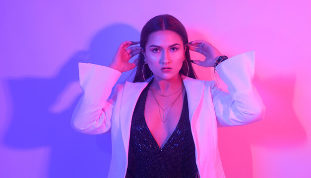 Portrait of a gorgeous brunette with big lips, perfect eyebrows, in big earrings, ringlets. A chic girl in a neon light. A party. Girl in a white jacket.