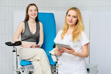 a young female gynecologist doctor with a tablet in her hands against the background of a female patient sitting in a blue gynecological chair and a colposcope. Women's consultation