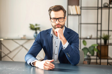 Portrait of sophisticated young bearded confident entrepreneur man in business casual clothes sitting at the desk and looking at camera, placed hand on his chin and thinking. Copy space concept