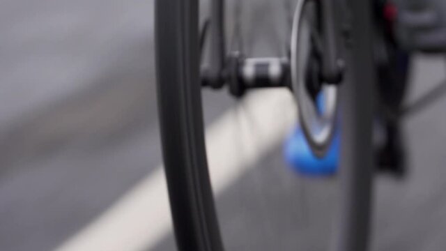 Close Up Clip Of Narrow Front Wheel Of A Road Bike, Showing Grip And Tread Of The Tire And Rotation, With Blue Cycling Shoes And White Road Markings In The Background