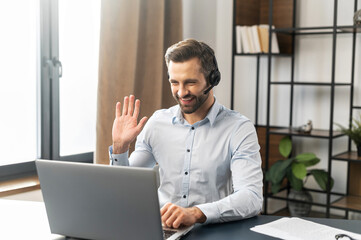 Fototapeta premium Portrait of fit handsome young businessman in casual shirt and headset looking on laptop, customer service representative sitting at the home office and greeting client, waving, saying hello, typing