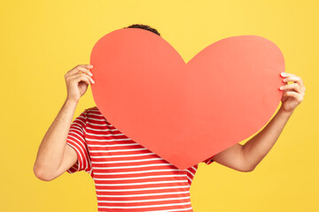 Unknown man in red striped t-shirt hiding his face behind big red paper heart, making anonymous surprise, sending greeting card. Indoor studio shot isolated on yellow background