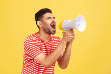 Angry nervous man with beard in striped t-shirt loudly screaming at megaphone, making announce, protesting, wants to be heard. Indoor studio shot isolated on yellow background