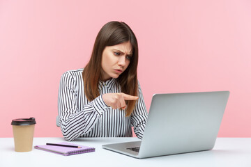 Hey you! Angry business woman in striped shirt pointing to laptop screen, scolding and accusing talking on video call, having online conference. Indoor studio shot isolated on pink background
