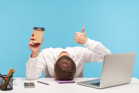 Tired Man Office Worker Lying Face Down On Desk Holding Paper Cup Of Coffee And Showing Thumbs Up, Need Coffee To Be Ok, Overworking. Indoor Studio Shot Isolated On Blue Background