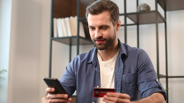 Young bearded hipster guy in casual clothes using mobile phone and credit card for online shopping, entering number to make payment, buying items, holding the debit card for internet banking account