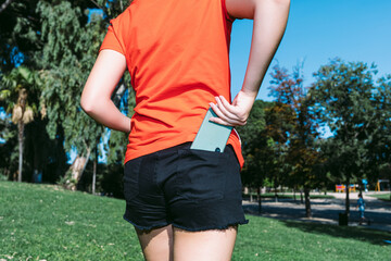 Young blonde woman in a red shirt keeps the mobile phone in the back pocket of her pants in a park