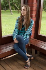 A beautiful young long-haired sad lonely girl in a plaid shirt sits on a park bench in summer or spring.