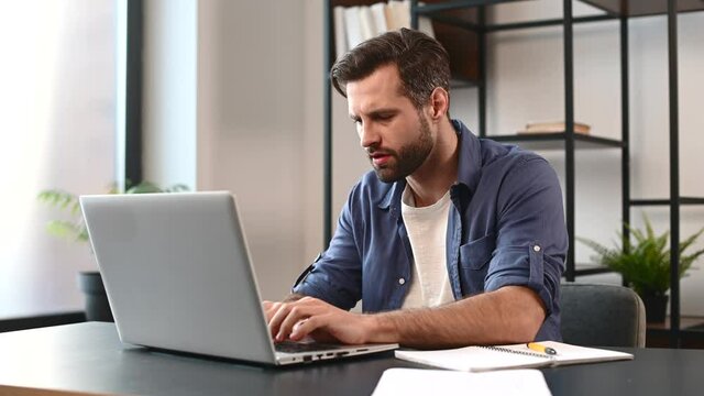 Young tired bearded office worker freelancer in casual clothes sitting at the desk at his workplace, supporting head with hand and looking at the laptop screen, feeling overworked and sleepy