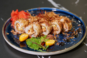 A close-up photo of sushi rolls with tuna shavings, Philadelphia cheese, and eel on a blue ceramic plate with sauce, kumquats, and leaves. Black stone table.