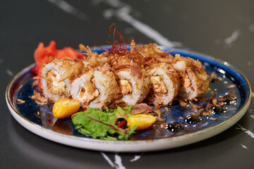 A close-up photo of sushi rolls with tuna shavings, Philadelphia cheese, and eel on a blue ceramic plate with sauce, lobules of kumquat, and leaves. Black stone table.