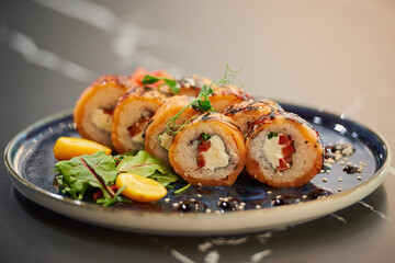 A close-up photo of sushi rolls with salmon, Philadelphia cheese, and tuna on a blue ceramic plate with sauce, sesame, lobules of kumquat, and leaves. Black stone table.