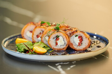 A close-up photo of fresh sushi rolls with salmon, Philadelphia cheese, and tuna on a blue ceramic plate with sauce, sesame, lobules of kumquat, and leaves. Black stone table.