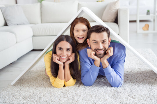 Photo of young happy cheerful smiling positive family lying on floor in new house relocation hold paper roof above heads
