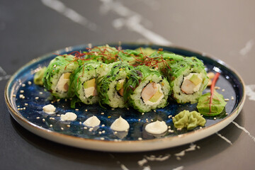 A close-up photo of sushi rolls with Chuka wakame seaweed, Philadelphia cheese, and shrimps on a blue ceramic plate with sauce, cashew, and wasabi on a black stone table.