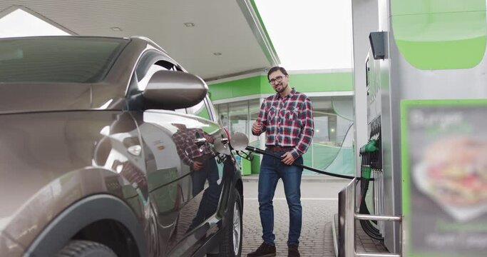 Portrait of a Young Handsome Bearded Caucasian Man with Glasses, He is standing at the gas station and holding a fuel injector in his hands. Man Smiling At Camera. The Driver Fills His Expensive Car