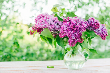 Fresh lilacs are arranged in a glass vase and placed on an outdoor table.