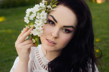 A girl sits on the grass in a park in a short white boho dress. The girl holds in her hands the first blossoms, a white branch of flowers. Brunette with long hair, spring has come. Gentle makeup