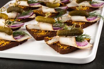 Blurred image of canapes with bacon, pickled cucumbers, dill and onions in a white plate against a black background.
