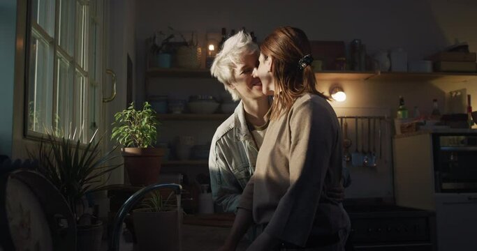 Cinematic shot of young carefree happy smiling homosexual female gay and transgender man couple in love is enjoying time together and kissing with affection while cooking in kitchen at home.
