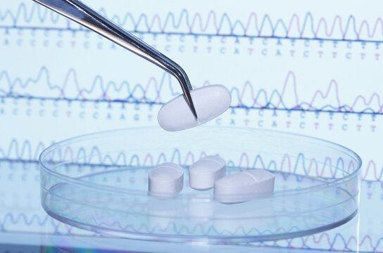 Tweezers holding pill over Petri dish. A computer screen that displays results of automated DNA sequencing is in the background