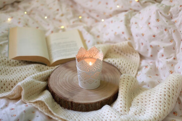 Candle holder with lit candle, wooden tray, open book and knitted blanket at home. Selective focus. 