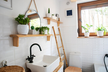 Bathroom with white tiles, window, stylish basin, round mirror, plants and wooden shelf in rustic...