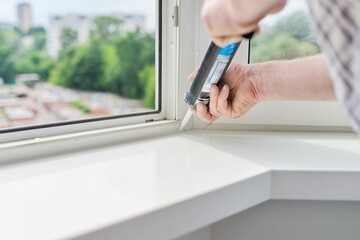 A worker with a construction syringe fills seam between sill and window with silicone sealant