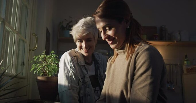 Cinematic shot of young carefree happy smiling homosexual female gay and transgender man couple in love is enjoying time together and kissing with affection while cooking in kitchen at home.