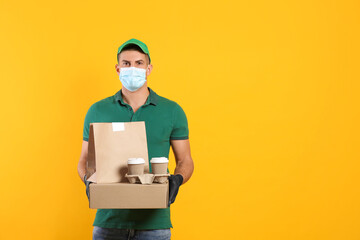 Fototapeta na wymiar Courier in medical mask holding packages with takeaway food and drinks on yellow background, space for text. Delivery service during quarantine due to Covid-19 outbreak