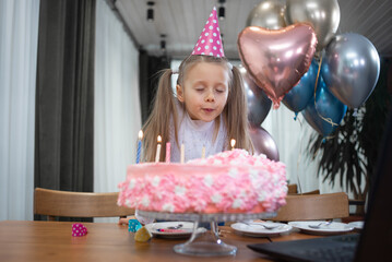 A girl celebrates her birthday via the Internet at quarantine time. blows out the candles on the birthday cake near the laptop. Festive decor, sweets, balloons. Daughters with parents children's party