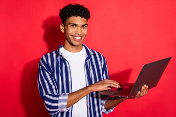 Portrait of attractive cheerful brunet guy holding in hands using laptop it project isolated over bright red color background