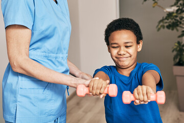 African American boy trains with physiotherapist using dumbbells at rehab center