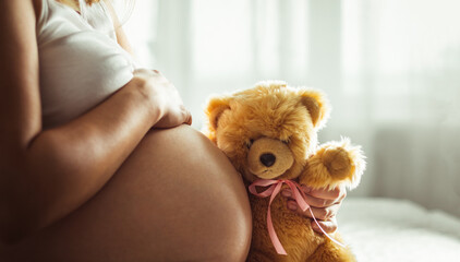 Pregnant woman touching her big naked belly and holding teddybear with pink ribbon. Baby girl expectation. Love and care. Close up