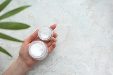 Cosmetic cream jars in hand. Woman holding white container with moisturizer marble background with copy space, defocused tropical leaf. Natural skin body or facial care concept