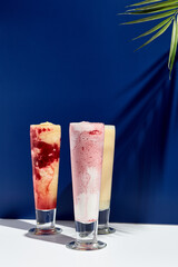 Raspberry and banana, pineapple, strawberry and coconut milk smoothie over white and blue background. Palm leaf with sunshine and hard shadow. Summer, healthy, fresh drink concept
