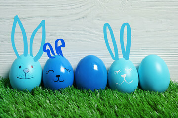 Several eggs with drawn faces and ears as Easter bunnies among others on green grass against white wooden background