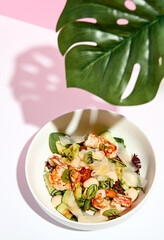 Poke bowl with shrimp and vegetables. Restaurant seafood appetizer on white table with pink wall. Day sunlight with hard shadow of monstera palm leaves. Summer or spring restaurant food concept.