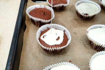 Chocolate brown muffin cake wrapped in white paper and covered with white frosting, baked in the oven, lying on baking paper.