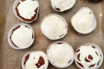 Chocolate brown muffin cake wrapped in white paper and covered with white frosting, baked in the oven, lying on baking paper, top view.