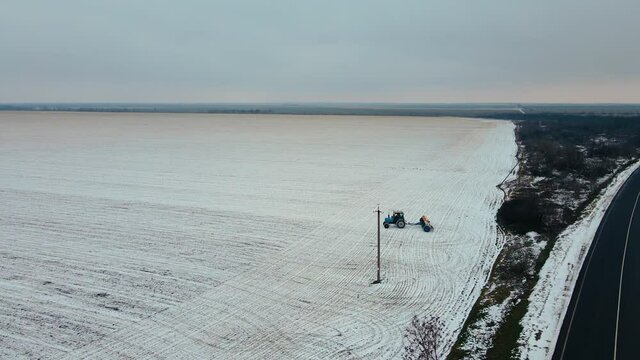 Drone shoots video of agricultural farmer on tractor with fertilizer adorable crops in winter. Snowy frozen highway
